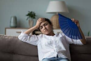 Woman fanning herself while leaning on a sofa, looking overheated and unwell, symbolizing hormonal imbalance and discomfort from hot indoor weather