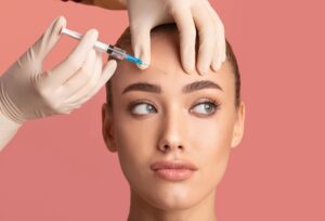Young woman receiving a Botox beauty injection in her forehead for skin lifting and wrinkle reduction, standing against a pink studio background